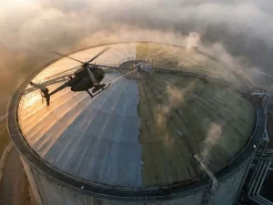 Helicopter scanning a circular biogas membrane dome at sunrise, laser beam cutting through morning haze over half-surveyed facility.