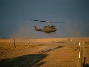 Helicopter patrolling buried gas pipeline route over flat farmland at golden hour, with warning posts and a hairline ground crack visible below.