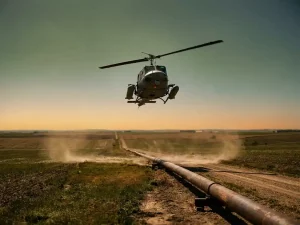 Helicopter with laser sensors flying low over a rural gas pipeline corridor, scanning farmland beneath an amber afternoon sky.