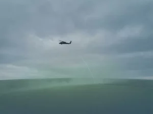 Helicopter silhouette flying low over a flat landscape with a laser beam arc and subtle green atmospheric haze below.