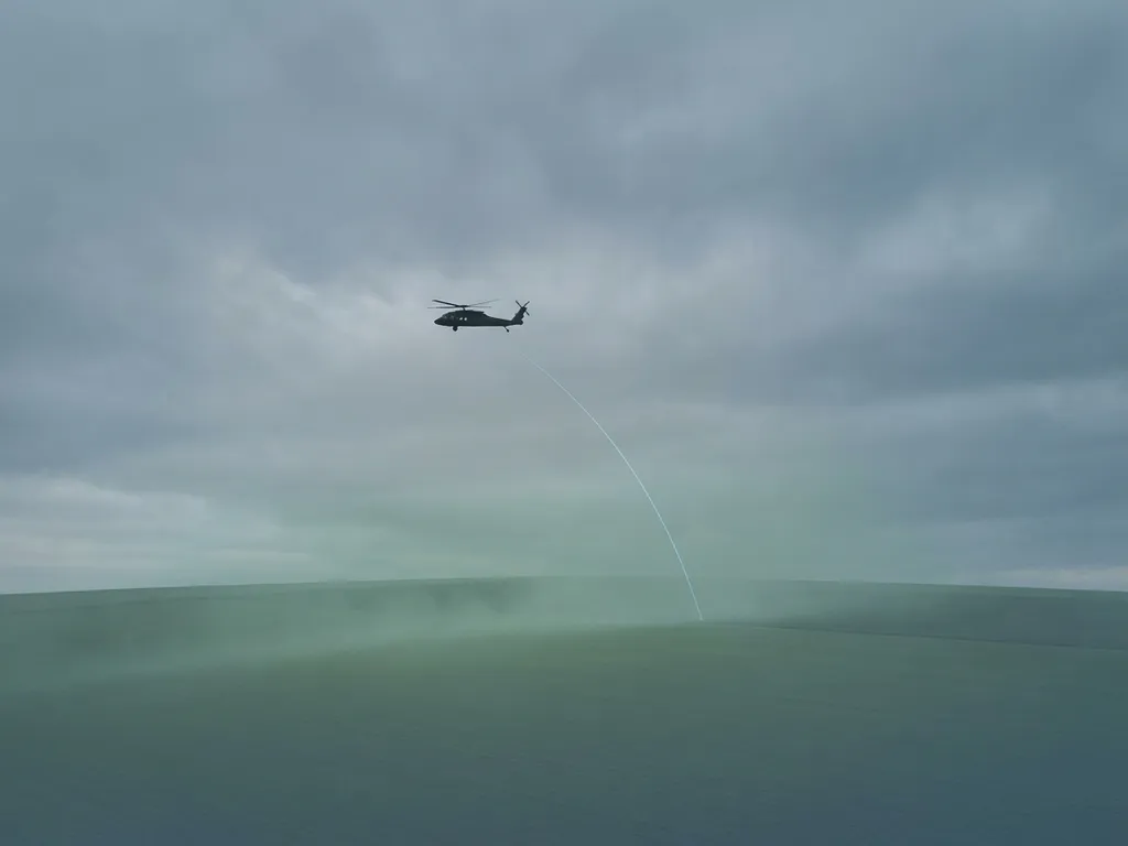 Helicopter silhouette flying low over a flat landscape with a laser beam arc and subtle green atmospheric haze below.