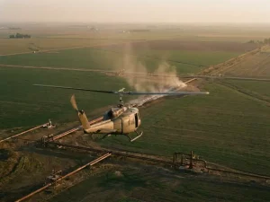 Helicopter flying low over green agricultural fields and industrial pipeline infrastructure during golden hour, aerial perspective.