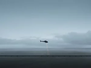 Helicopter silhouette flying low over a dark pipeline, projecting two amber laser beams onto a misty flat landscape under overcast sky.