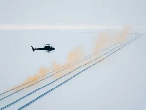 Helicopter flying low over parallel oil pipelines with faint amber methane haze rising from the ground under pale diffused daylight.