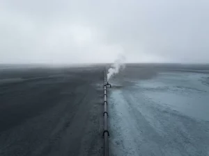 Aerial view of a solitary industrial pipeline crossing a flat landscape with a single pale gas plume rising against an overcast grey sky.