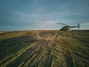 Helicopter conducting low-altitude methane survey over a cracked landfill cover at dusk, golden light casting long shadows across dusty terrain.