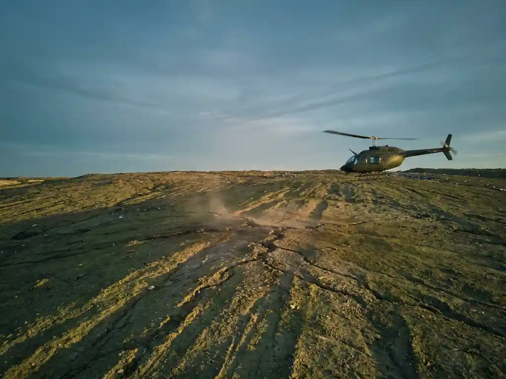 Helicopter conducting low-altitude methane survey over a cracked landfill cover at dusk, golden light casting long shadows across dusty terrain.