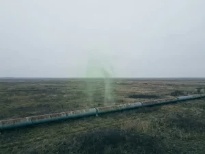 Weathered steel pipeline stretching to the horizon across a flat European landscape with a faint translucent green methane plume rising above it.