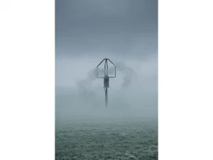 Methane sensor probe rising through dense morning fog above an open field, steel-grey stormy sky, minimalist low-angle composition.