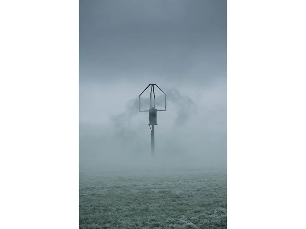 Methane sensor probe rising through dense morning fog above an open field, steel-grey stormy sky, minimalist low-angle composition.