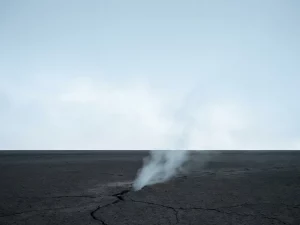 Pale vapor rising from cracked dark earth against a vast arctic blue sky, minimalist composition with geometric horizon line.