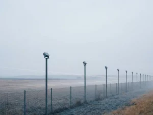 Slim geometric sensor poles along an industrial perimeter fence with faint laser beams in misty morning light.
