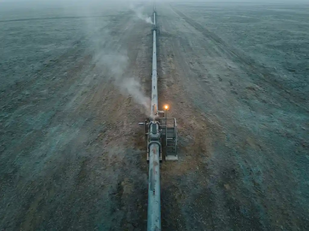 Industrial gas pipeline crossing a flat landscape with faint translucent plumes rising into an overcast pale sky, aerial view.
