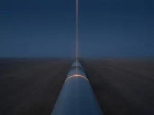 Steel pipeline stretching to a vanishing point across a flat landscape, scanned by an amber light beam under a deep midnight blue sky.