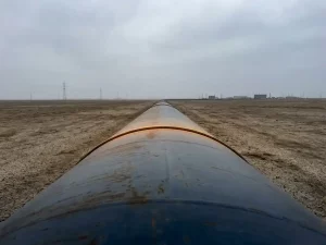 Large-diameter steel pipeline extending across a flat plain under overcast skies, shot at low eye level with amber light tracing its curve.
