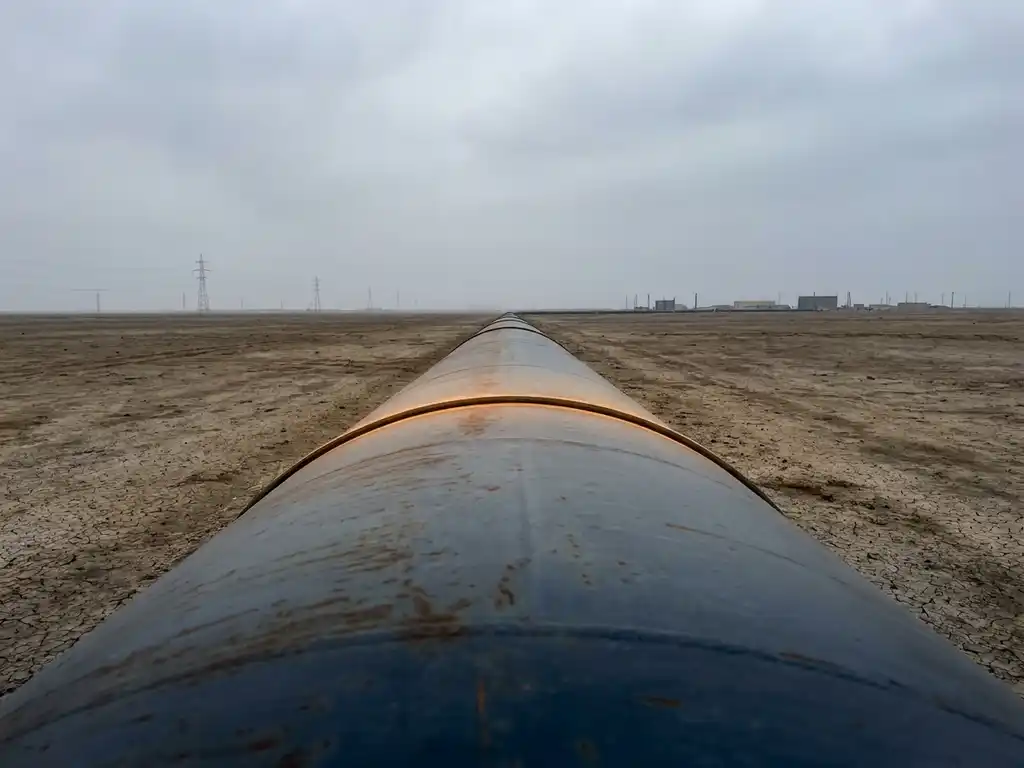 Large-diameter steel pipeline extending across a flat plain under overcast skies, shot at low eye level with amber light tracing its curve.