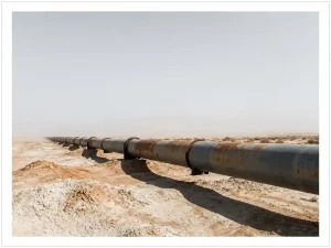 Weathered rust-streaked steel pipeline stretching across dry cracked earth under a pale sky, shot from extreme low angle.